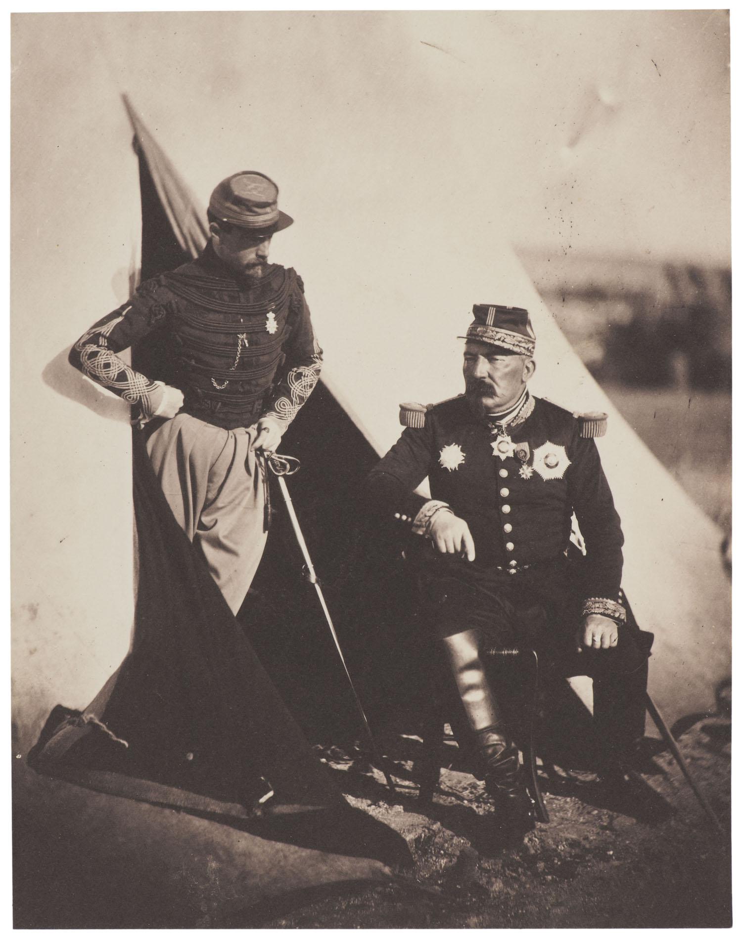 One man standing in the doorway of a tent, leaning on a rifle, another man sitting outside the tent. Both are wearing military uniforms.