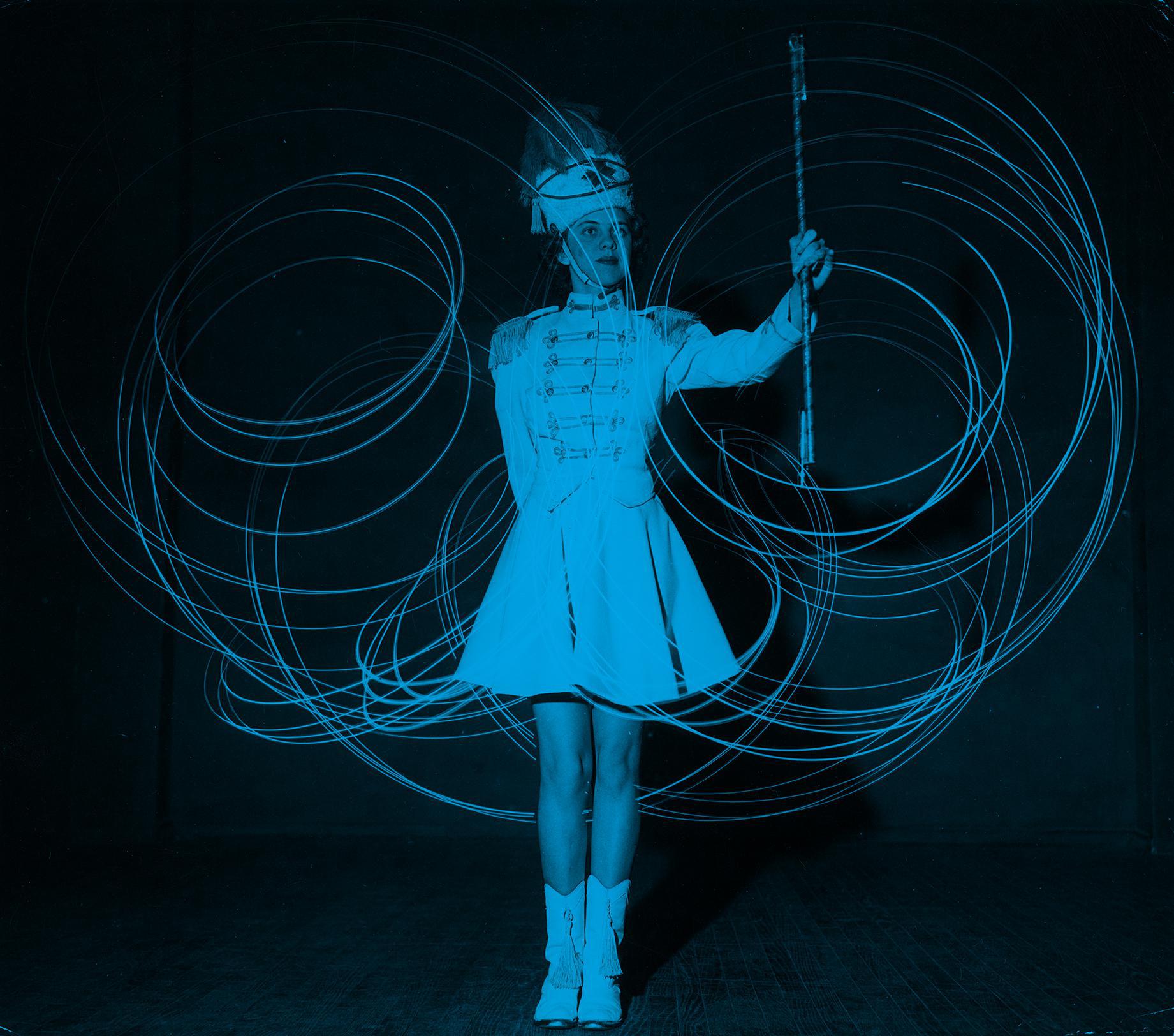 A woman in a white dress swirls a baton, leaving blue circles in its path
