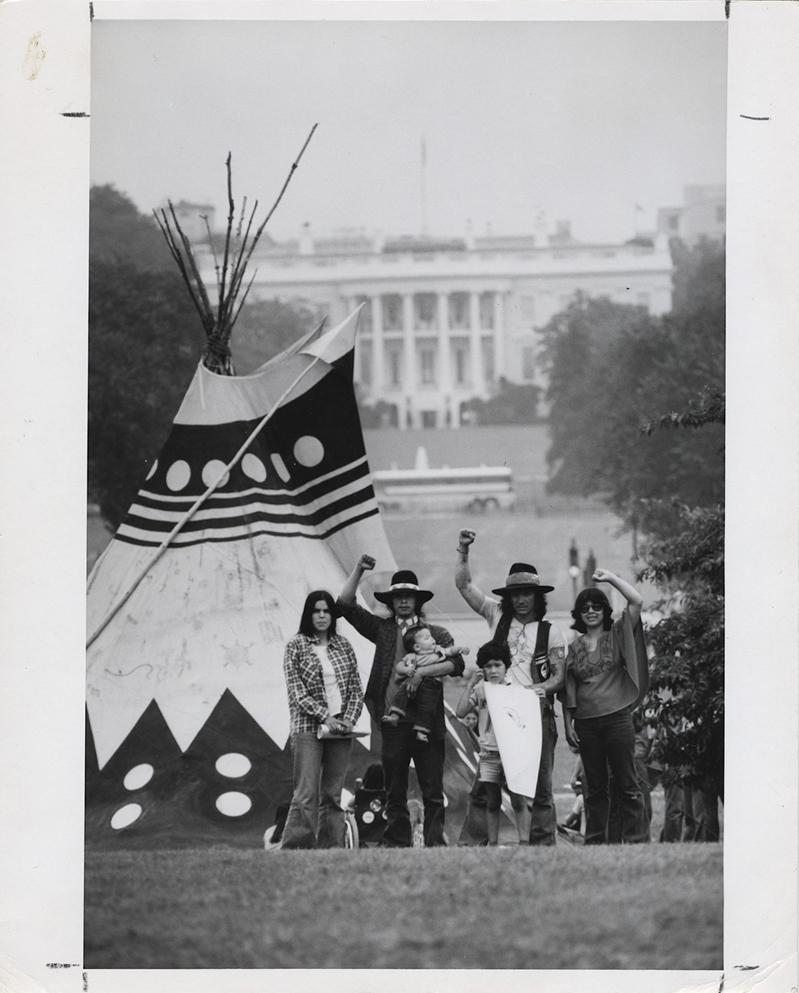 Four men in front of a tipi with their arms raised