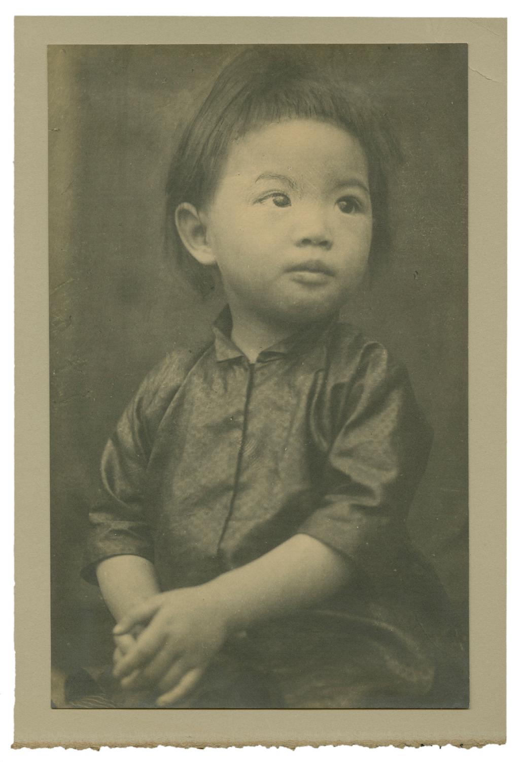 Studio portrait of a child facing right, looking up, hands clasped. Black and white photograph by Minna Keene.