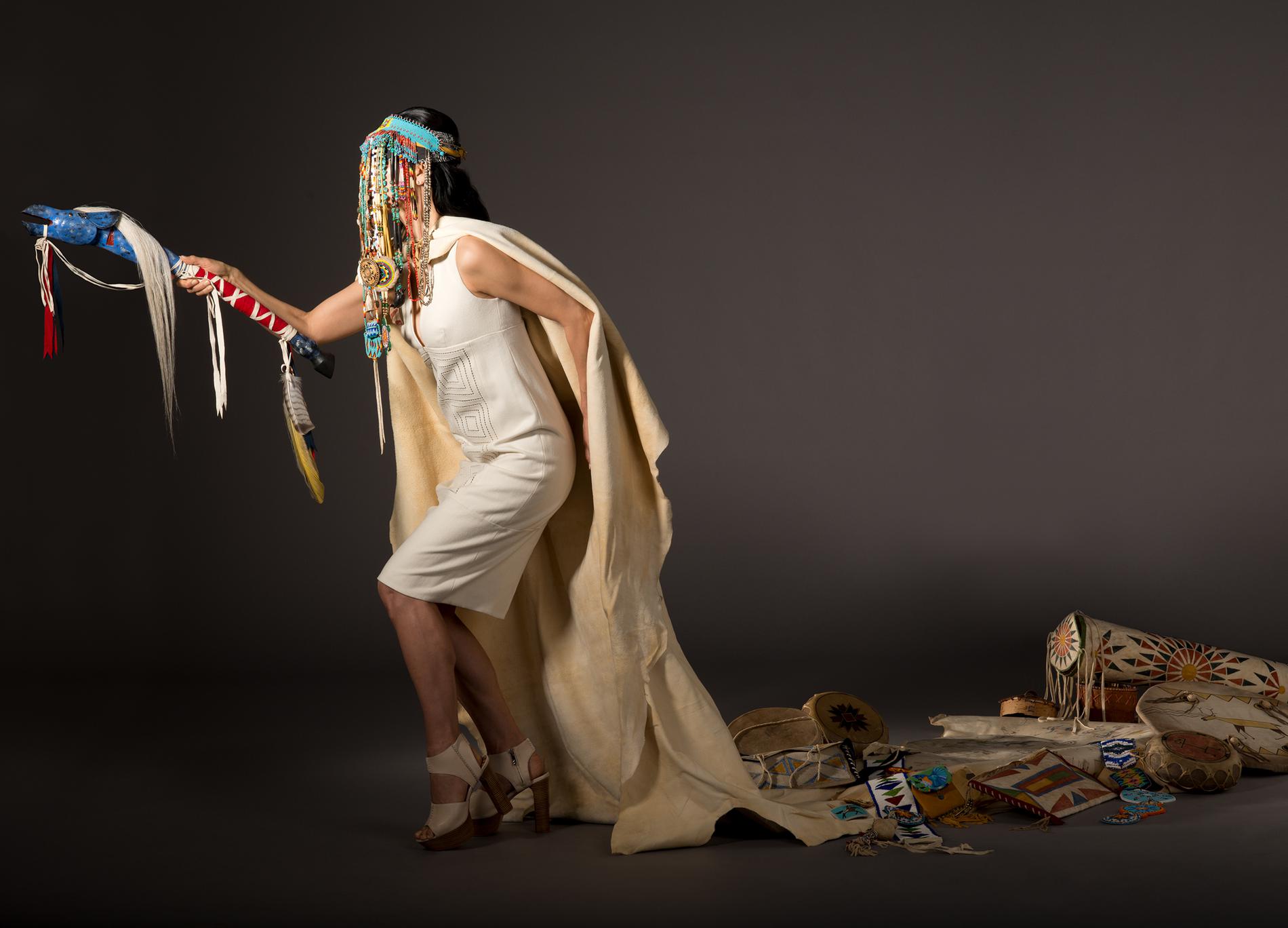 Indigenous woman with veiled beaded headdress and cape walks forward, carrying a multi-colourred staff with her arm outstretched, leaving a trail of Indigenous artifacts trailing on the floor behind her