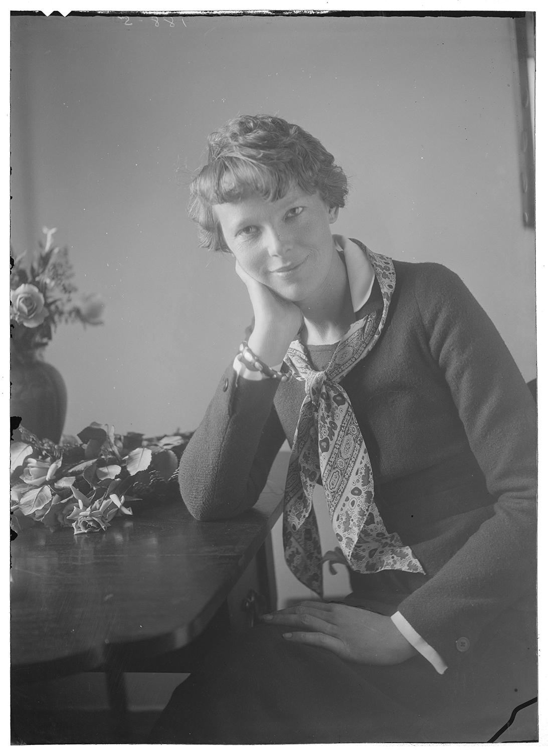 A portrait of a woman sitting down with her hand on her cheek and the other on her lap looking towards the camera. There is a vase of flowers beside her.  Black and white photograph by Violet Keene Perinchief.