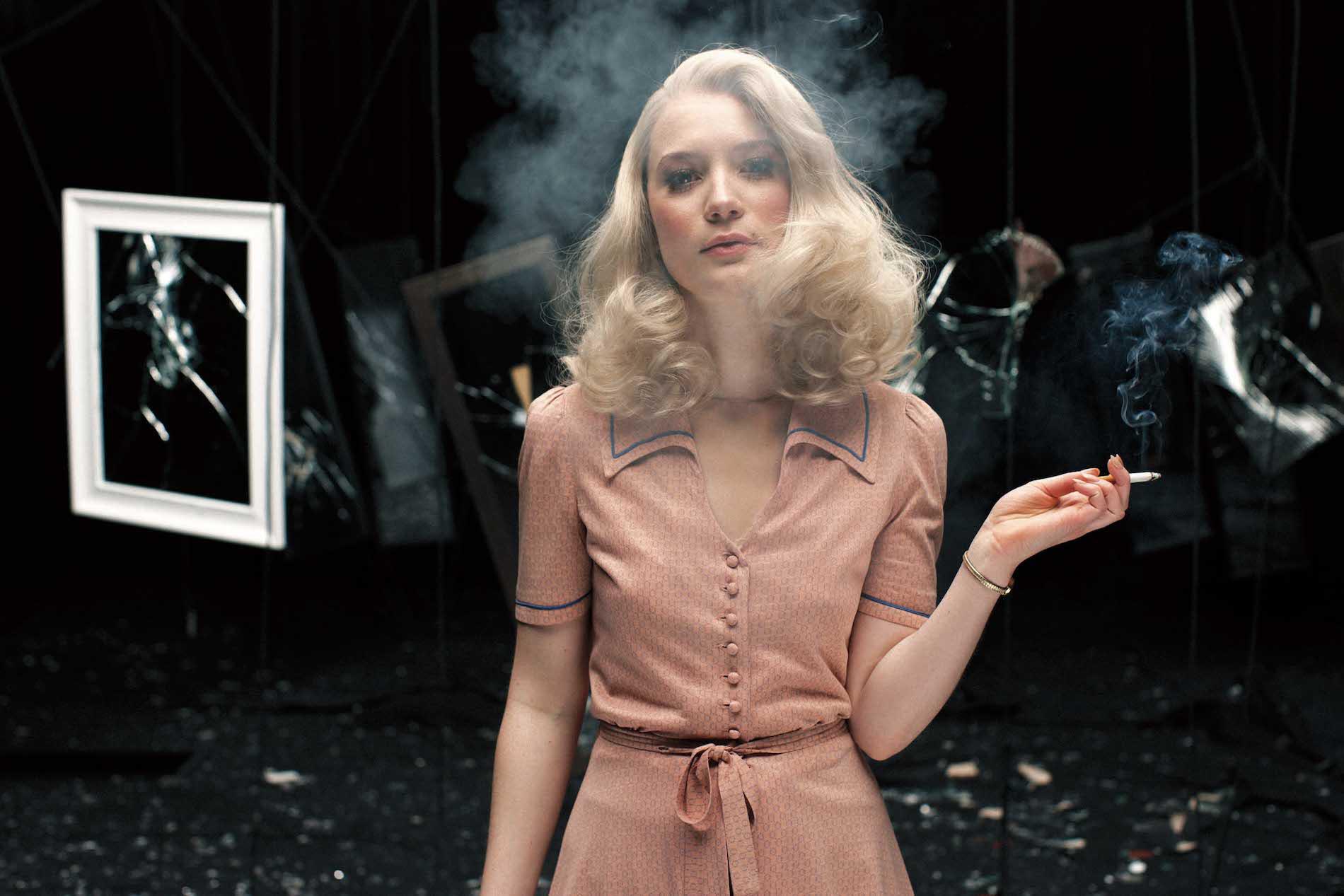 A blonde woman wearing a pink dress smoking a cigarette, smoke covering her face