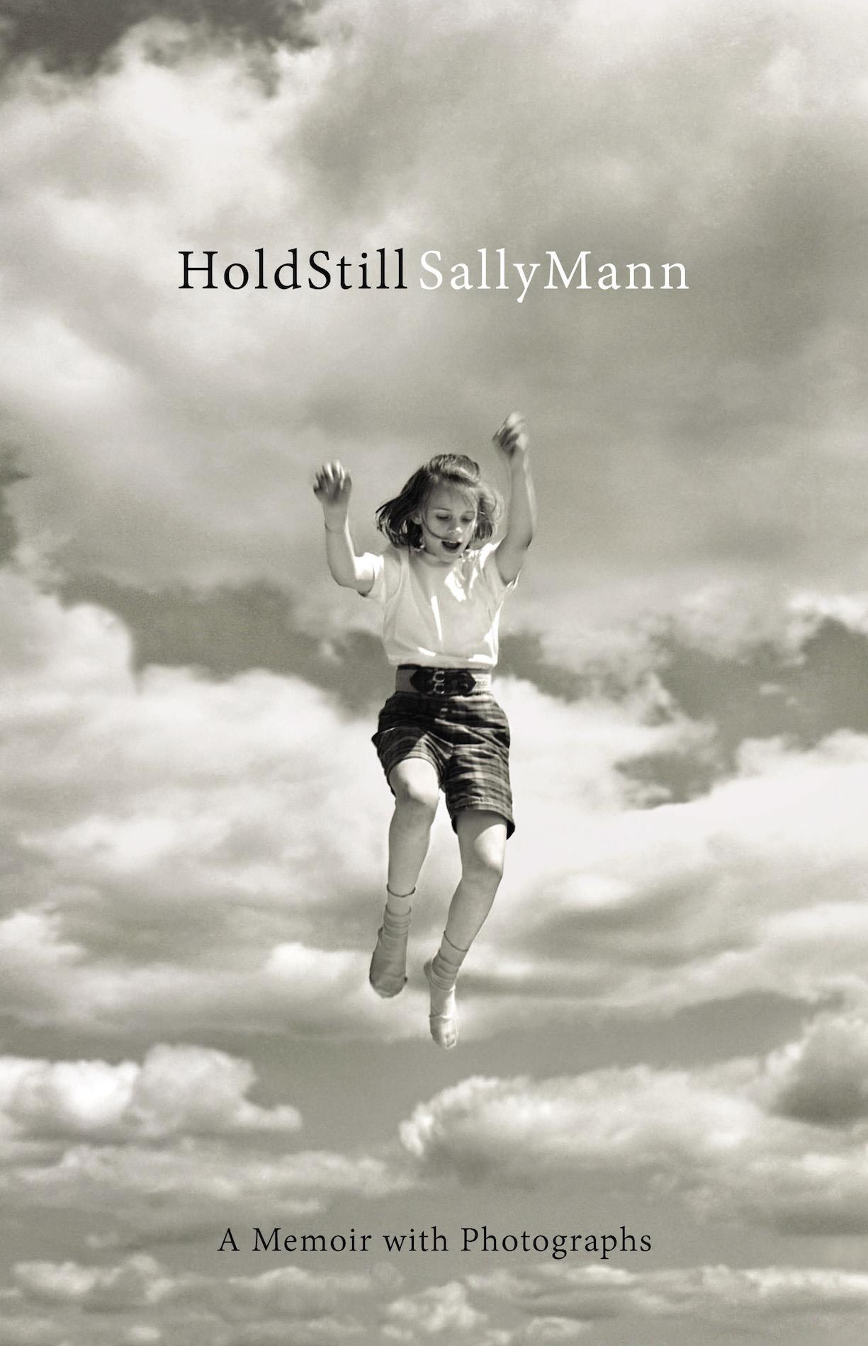 A book cover with an image of a girl wearing a tshirt, shorts, and long socks jumping. Text reads 'Hold Still. Sally Mann. A Memoir With Photographs'