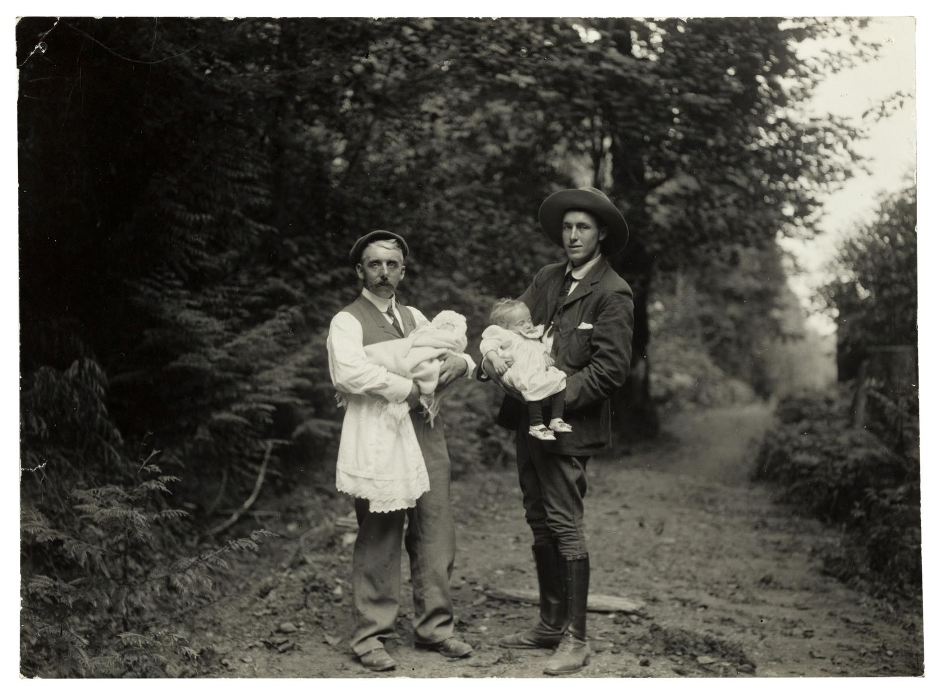 Two men holding babies on a forested path