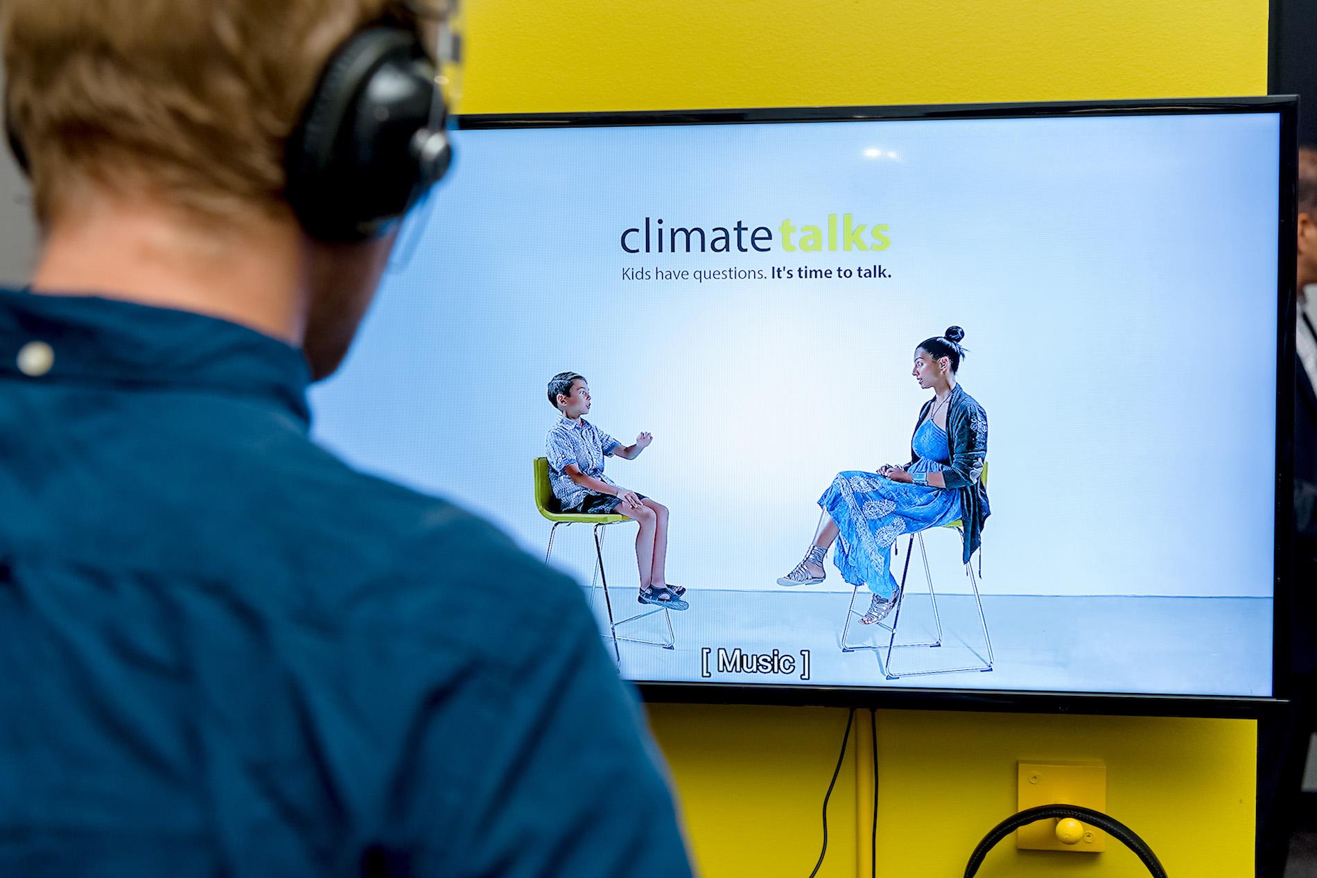 A man in a blue shirt watches a video on a screen, mounted on a yellow wall. On-screen text reads 'Climate Talks. Kids have questions. It's Time to Talk'