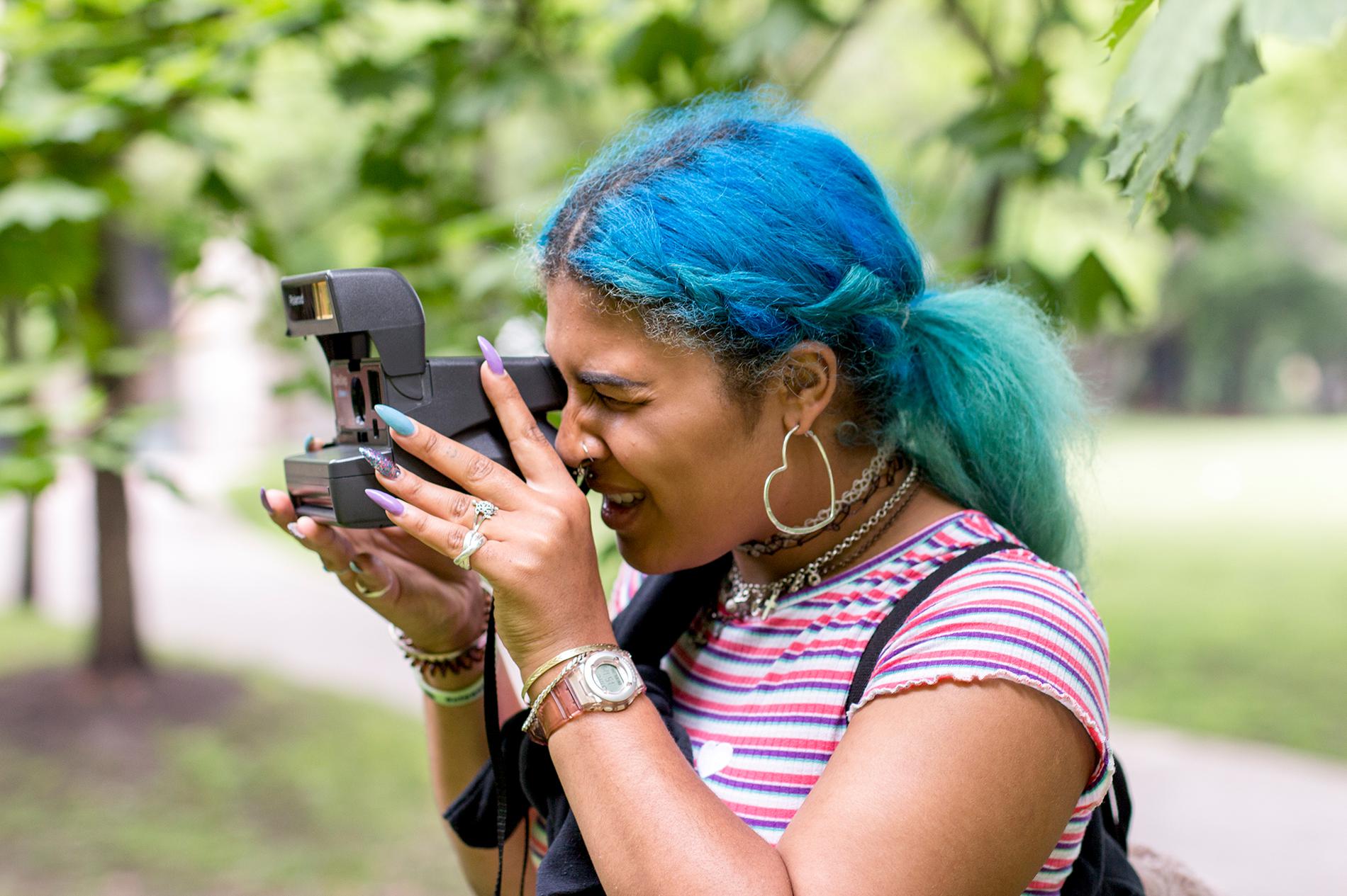 A young woman with blue hair taking a photo with a polaroid camera outside