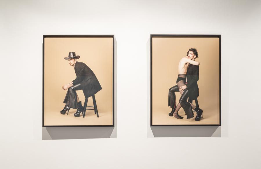 Two photographs on a wall: 1 with a man posing on a stool with high heels, and another with that same man and another man straddling him