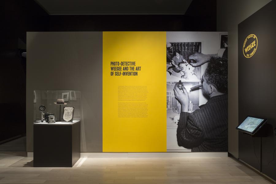 A glass display case with a camera and hat, a yellow wall with text that reads "photo-detective Weegee and the art of self invention", and a photo of Weegee marking up a photograph
