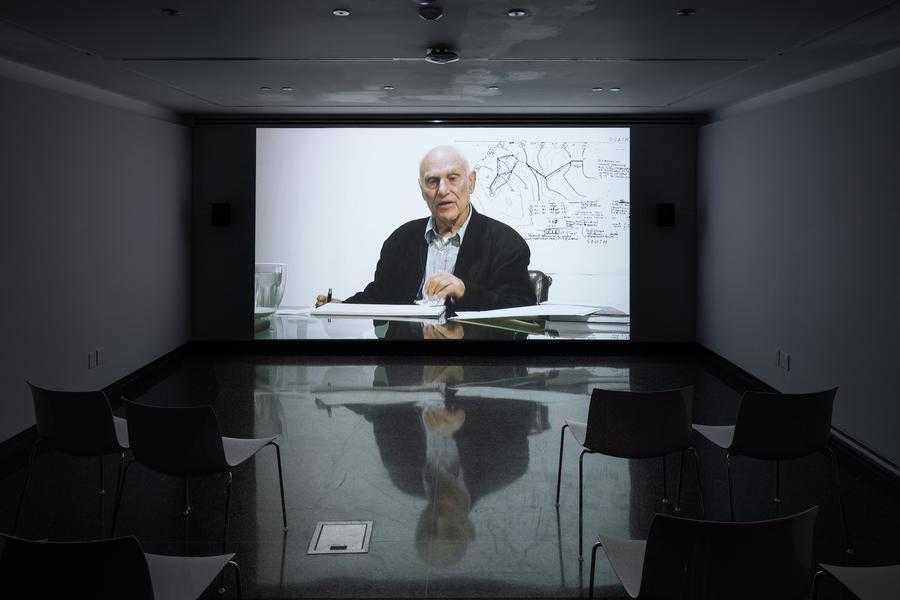 A projected video still of a man sitting at a desk speaking