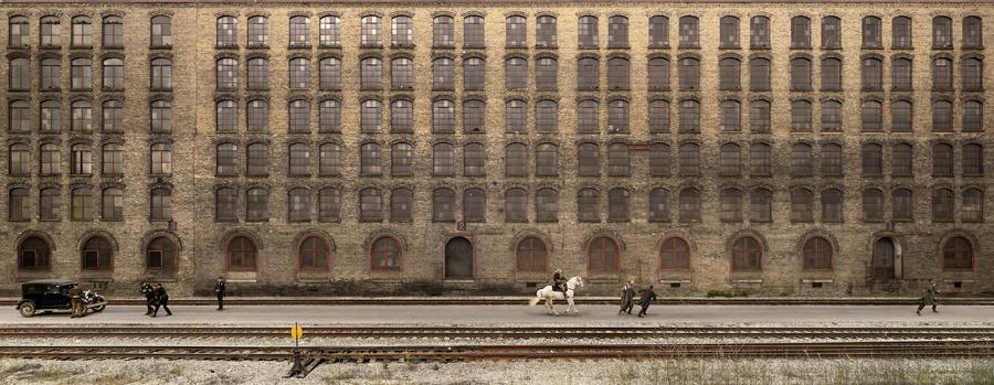 A wide shot of an old building, train tracks in the foreground. A group of men pull a white horse, another group pulls a black car