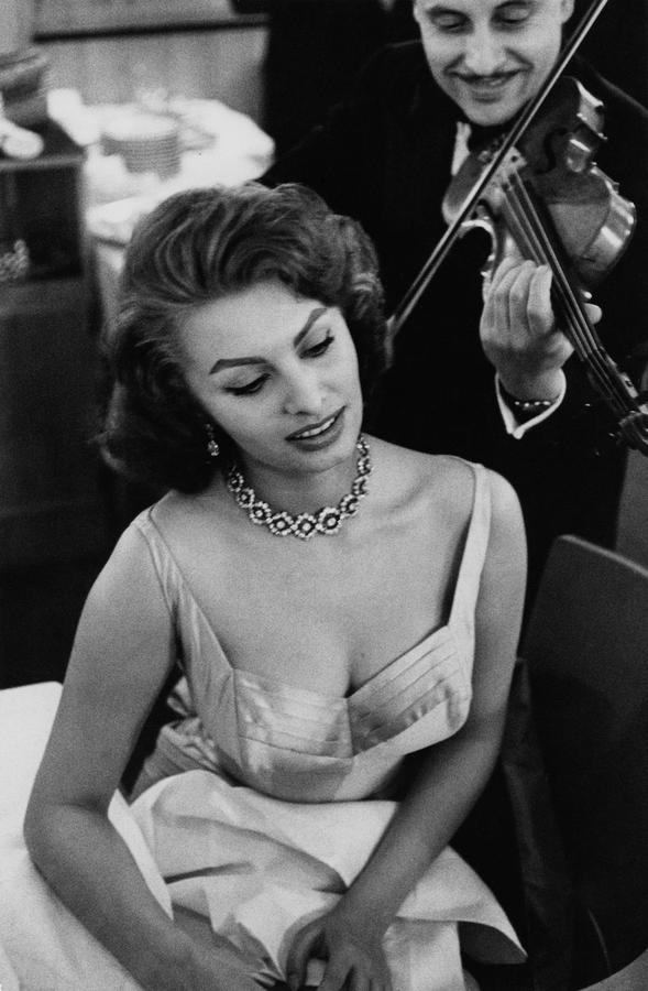 Sophia Loren wearing a formal dress and necklace, a man plays a violin behind her
