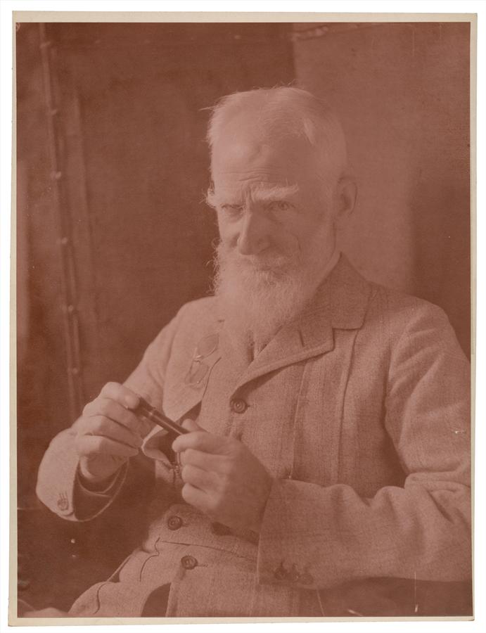 A studio portrait of a bearded man looking into the camera, holding a small cylinder object in his hand. Toned gelatin silver print by Violet Keene Perinchief.