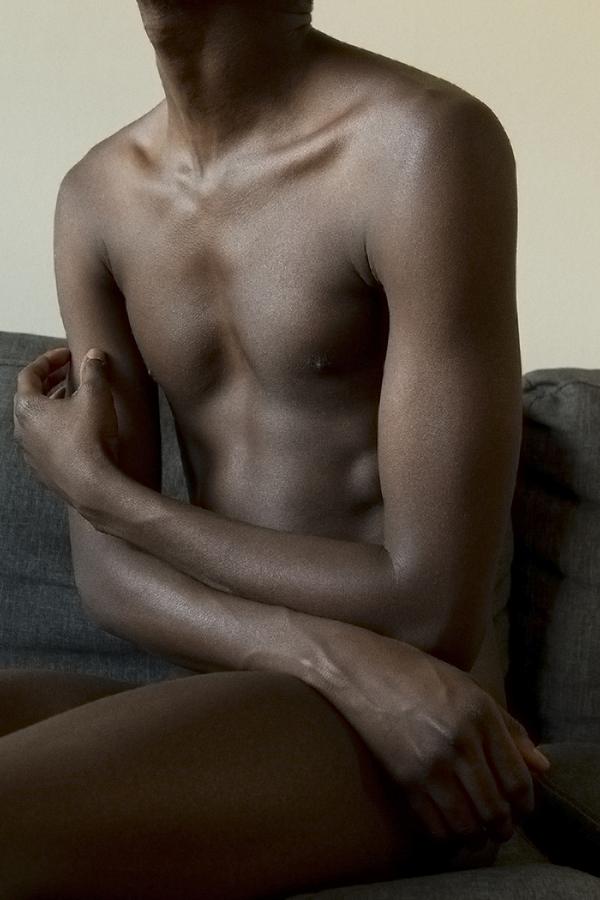 A naked man sits on a grey couch, arms loosely crossed
