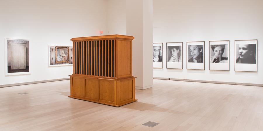 A wooden storage unit in the middle of the gallery, framed portraits on the wall behind it