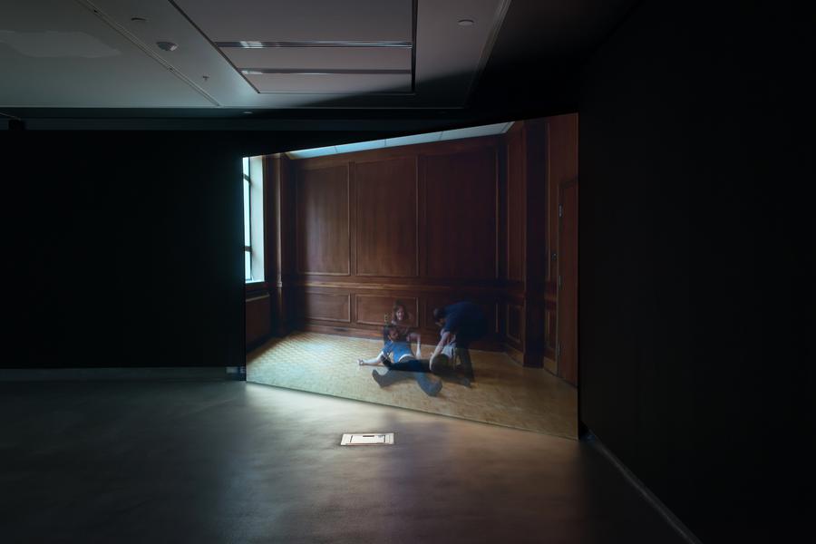 Video still of four people holding each other in the middle of a large empty room