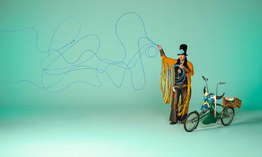A person throwing a lasso with a tall hat and a fringed jacket stands next to a low-rider bicycle in front of a turquoise background.