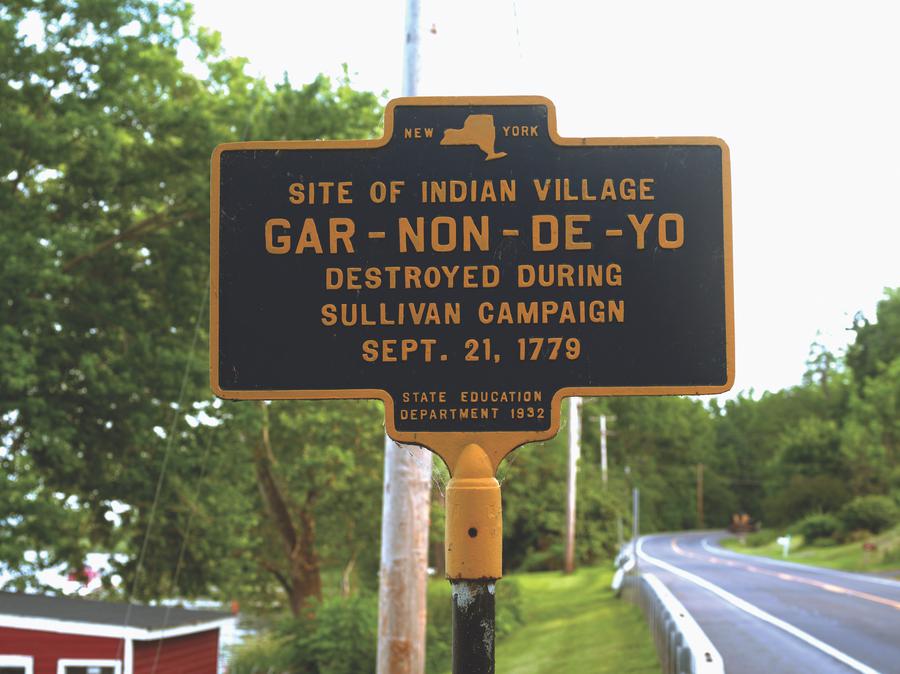 A black sign with yellow text that reads 'Site of Indian village. Gar-Non-De-Yo. Destroyed during Sullivan Campaign Sept. 21, 1779. State Education Department.'