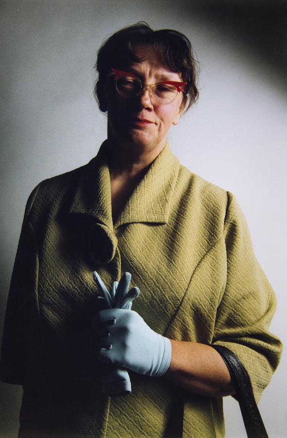 A woman wearing a green coat, red horn-rimmed glasses, and blue gloves