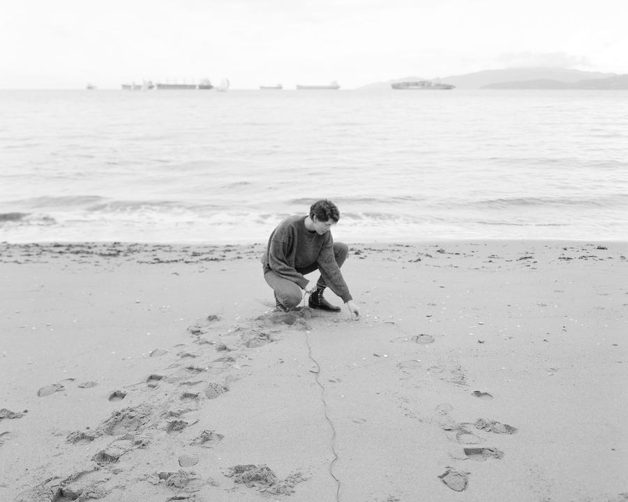 Person crouches down to touch sand on beach shore, cord extends from hand. Black and white