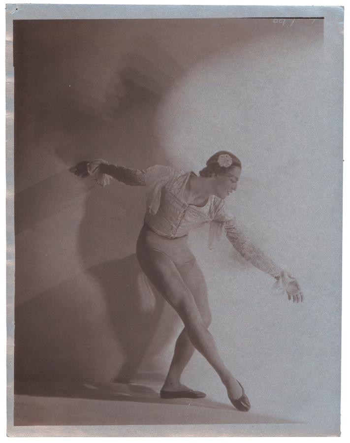 A dancer in front of a white wall doing a curtsey, peering towards the ground. Black and white photograph by Violet Keene Perinchief.