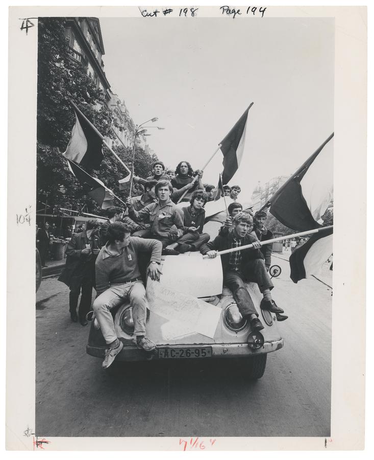 Young people carrying flags ride on top of a car in Prague.