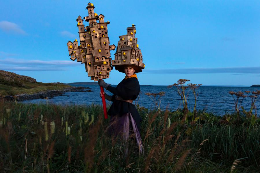 A woman wears on her head and holds in her hands two birdhouses, in front of a body of water