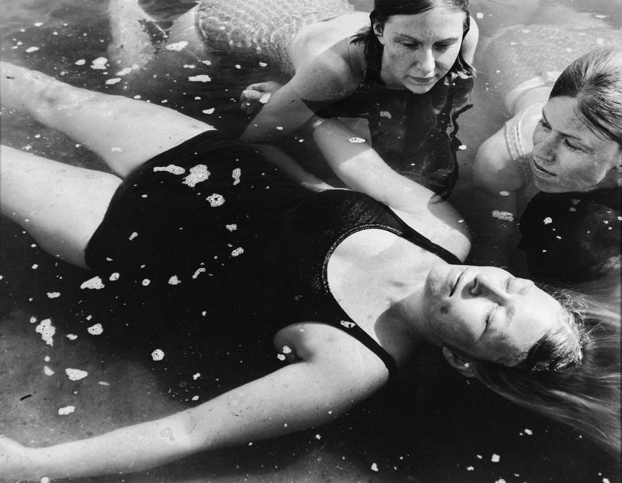A woman in a black swimsuit floats on her back with her eyes closed, two other women swimming watch
