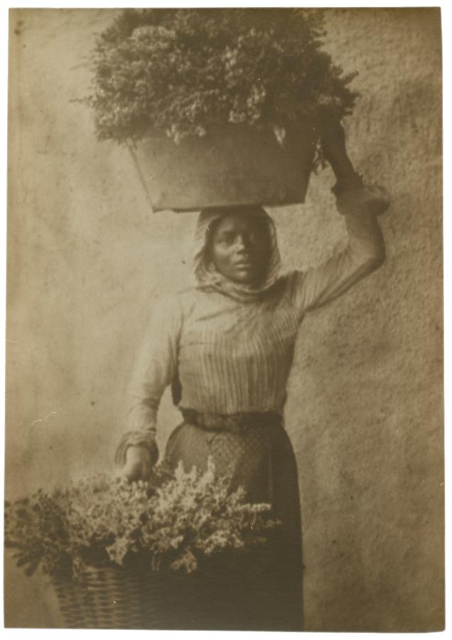 Portrait of a woman holding a large vessel with flowers on her head with her right hand and a large basket of flowers in her left hand by her waist. She is looking into the camera. Black and white photograph by Minna Keene.