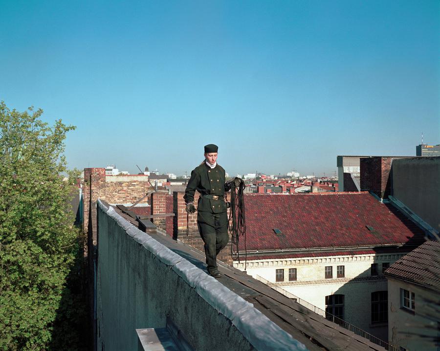 A woman in green uniform carries a rope across the rooftops of Berlin on a bright, sunny day.