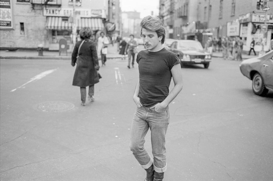 A black and white image of a man crossing an urban street with his hands in his pockets.