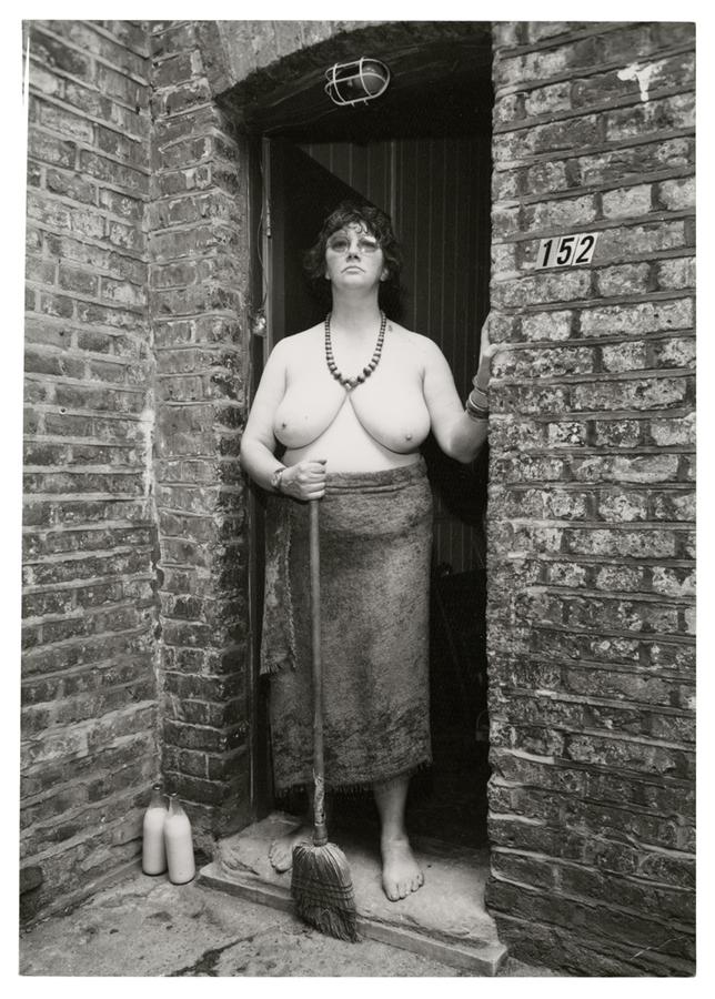 Photo of a woman with no shirt standing in a doorway holding a broom.