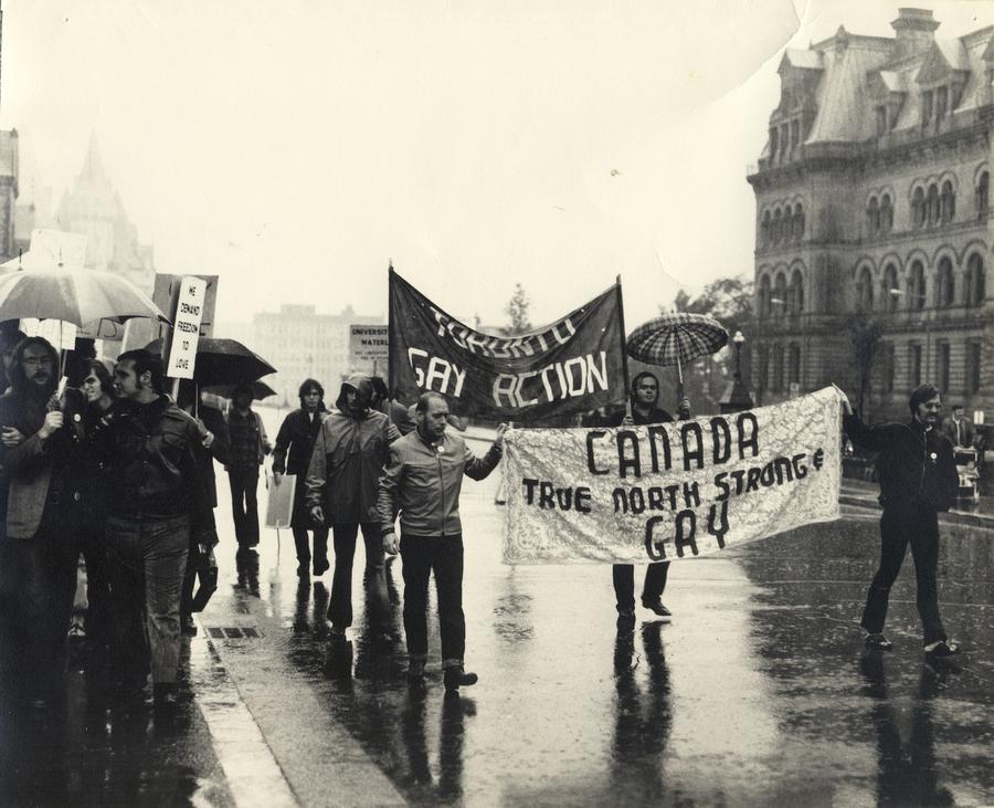 A rainy-day protest on Parliament Hill. Banners read "Canada true north strong & gay" and "Toronto gay action"