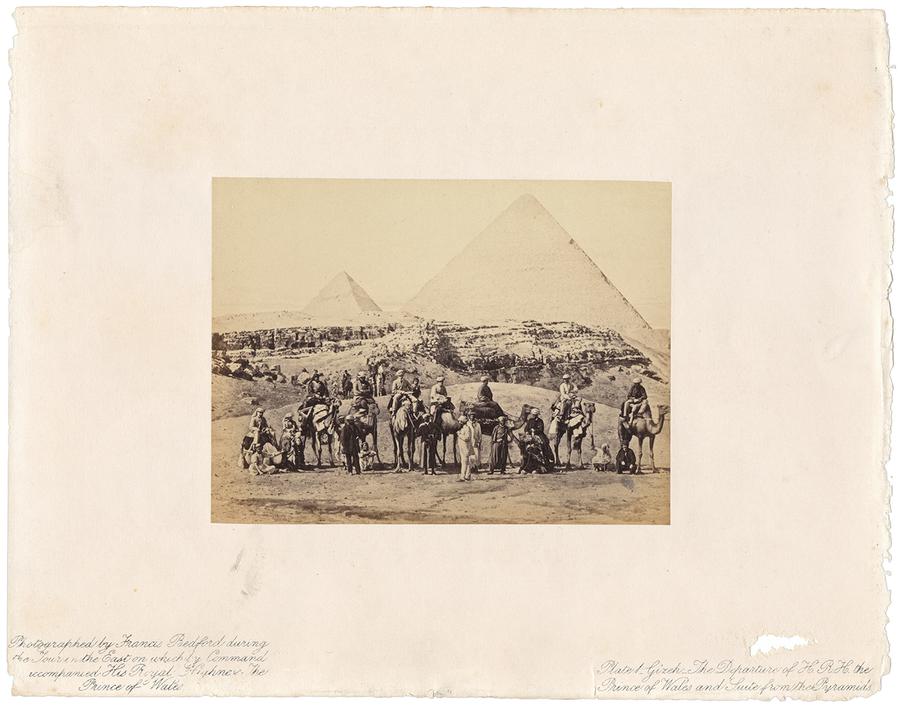 A group of men on camel pose in front of two pyramids in the background.