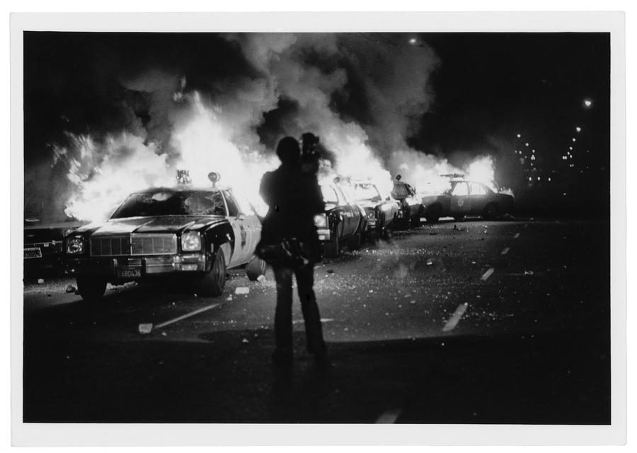 A group of police cars on fire, broken glass on the ground