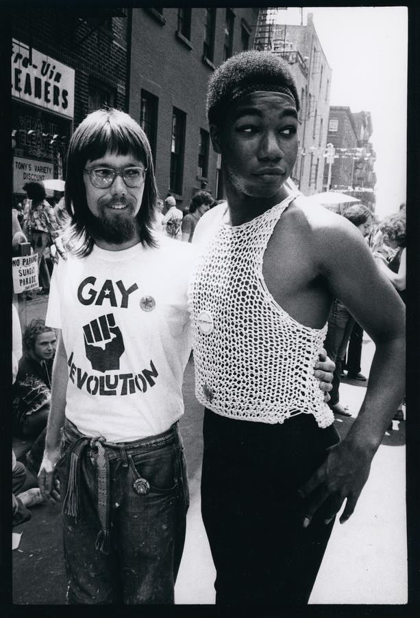 A man wearing a white shirt that says "gay revolution" has his arm around another man in  white mesh shirt