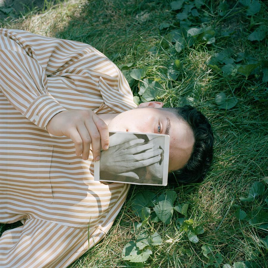 A person in a striped shirt lies on the grass; they hold a printed image of a hand covering one eye, while the other looks directly at the camera.