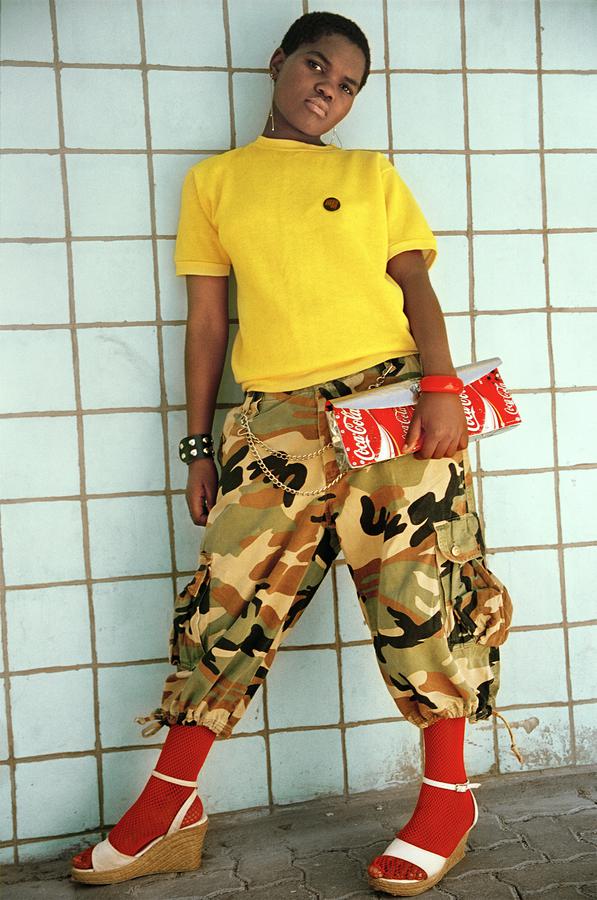 A young black woman in a yellow shirt, army pants, and heels, poses against a green wall, looking directly into the camera