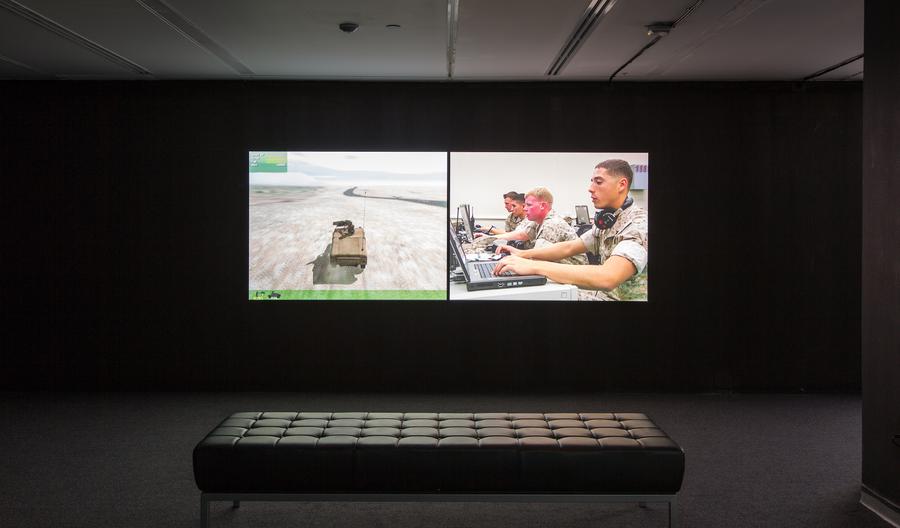 Two video stills in a dark room with a bench in the middle. On the left, a tank driving in the desert. On the right, four soldiers use computers