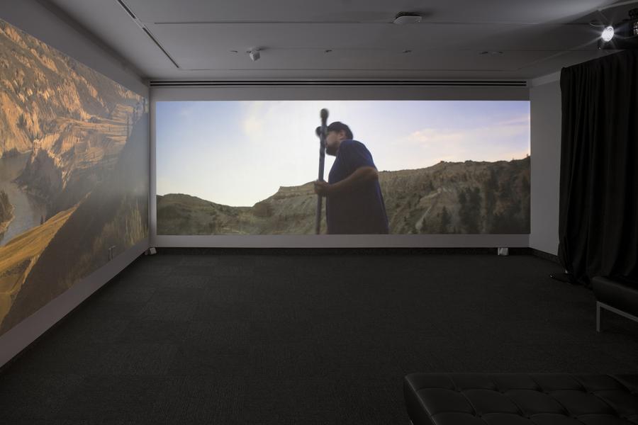 Two adjacent screens play video: on the left, a mountain range, on the right, a man stands with a tall stick. In the room, two black leather benches sit in the foreground