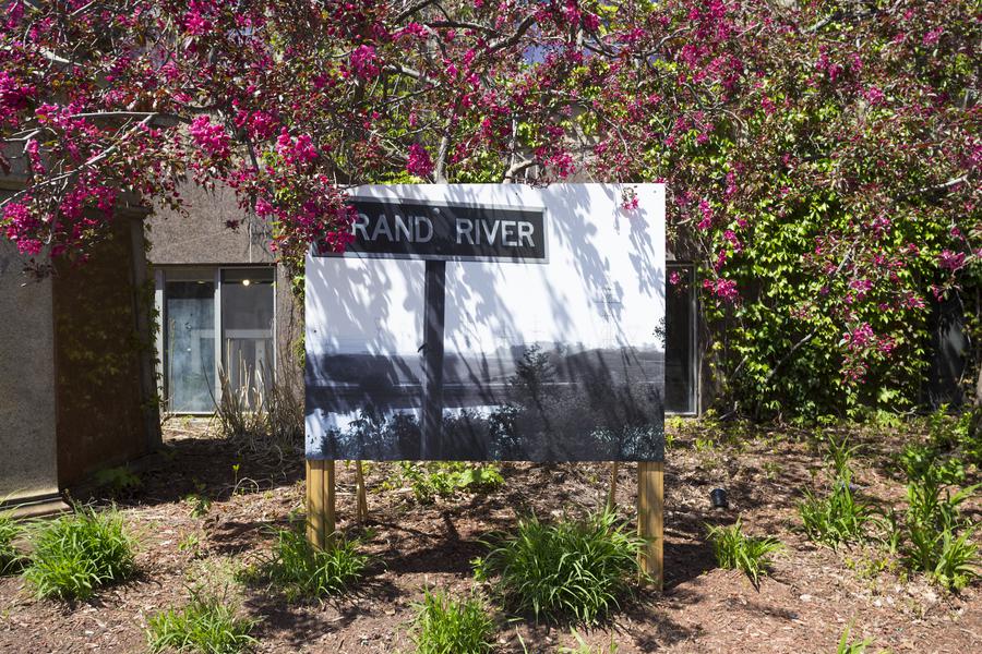 Black and white photograph of a road sign that reads 'Grand River', displayed outside under a tree with pink flowers