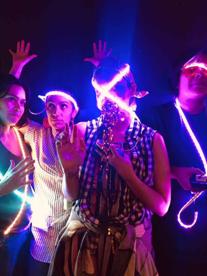 Four people standing in a dark room, draped in strands of neon lights