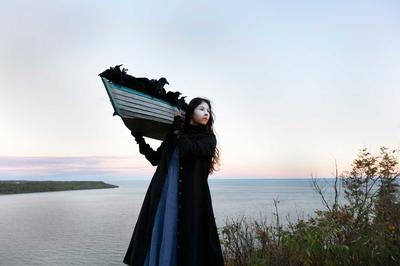 Overlooking a body of water, a woman with white face makeup wearing a black coat holds a small boat on her shoulder, filled with ravens