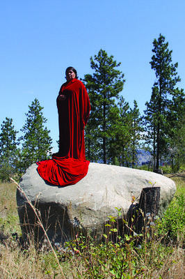 A woman dressed in a long red dress stands atop a large rock